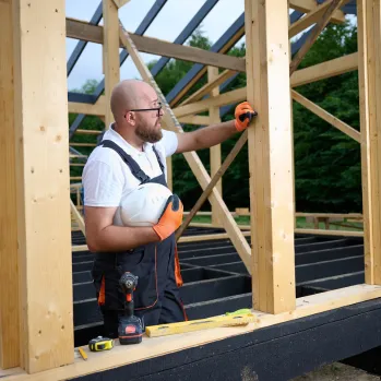 Contractor wearing a yellow hard hat and safety harness working on a wooden frame at a construction site.